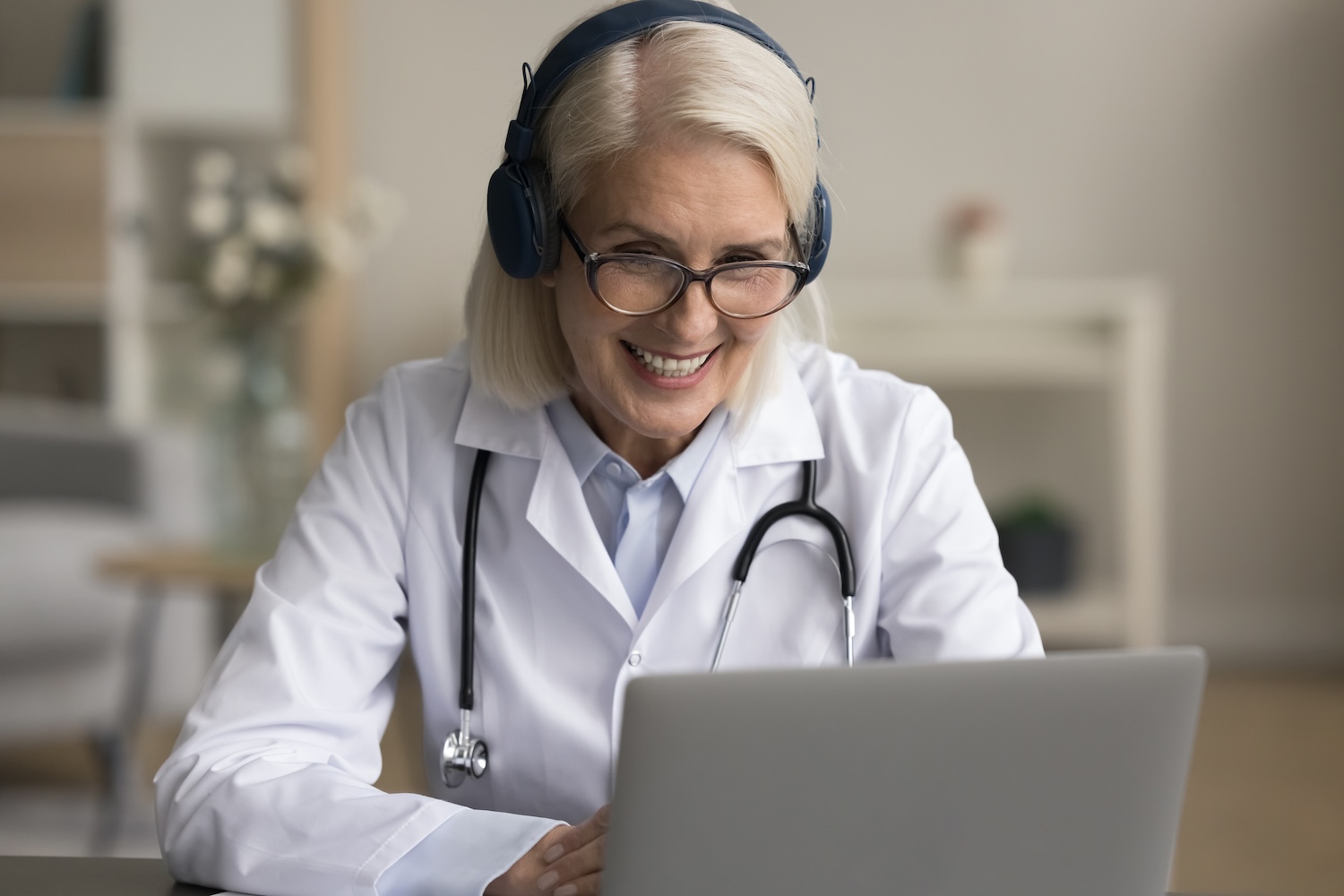 female in white lab coat with stethoscope around her neck and headset on smiling at laptop screen